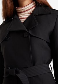 Žena v černém trench coatě páskovém, který má přes červený a bílý pruhovaný rolák, zobrazena od krku po pas.