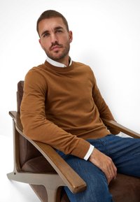 Bruine gebreide trui met een ronde halslijn, gedragen over een witte blouse, gecombineerd met blauwe spijkerbroek, zittend in een houten fauteuil.