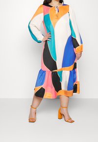 Femme portant une robe midi colorée avec des formes abstraites en orange, bleu, rose, noir, blanc et bleu sarcelle, associée à des sandales à talons blocs orange.