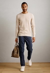 Άνδρας που περπατάει σε εσωτερικό χώρο φορώντας ένα μπεζ πουλόβερ με καλώδιο, ναυτικές παντελόνες, λευκά αθλητικά παπούτσια και κρατώντας μια καφέ δερμάτινη τσάντα στο αριστερό του χέρι.