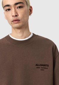 Sudadera marrón hecha de tela suave, con cuello redondo, puños acanalados y un logo discreto en el pecho en color negro.