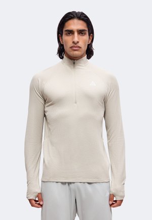 Uomo con i capelli scuri che indossa una maglia beige a maniche lunghe con cerniera e pantaloni grigio chiaro, in piedi davanti a uno sfondo grigio chiaro, rivolto verso l'obiettivo.