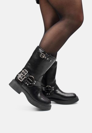 Piernas con medias negras translúcidas y botas de cuero negras hasta la mitad de la pantorrilla con múltiples hebillas plateadas y suelas gruesas sobre fondo claro.