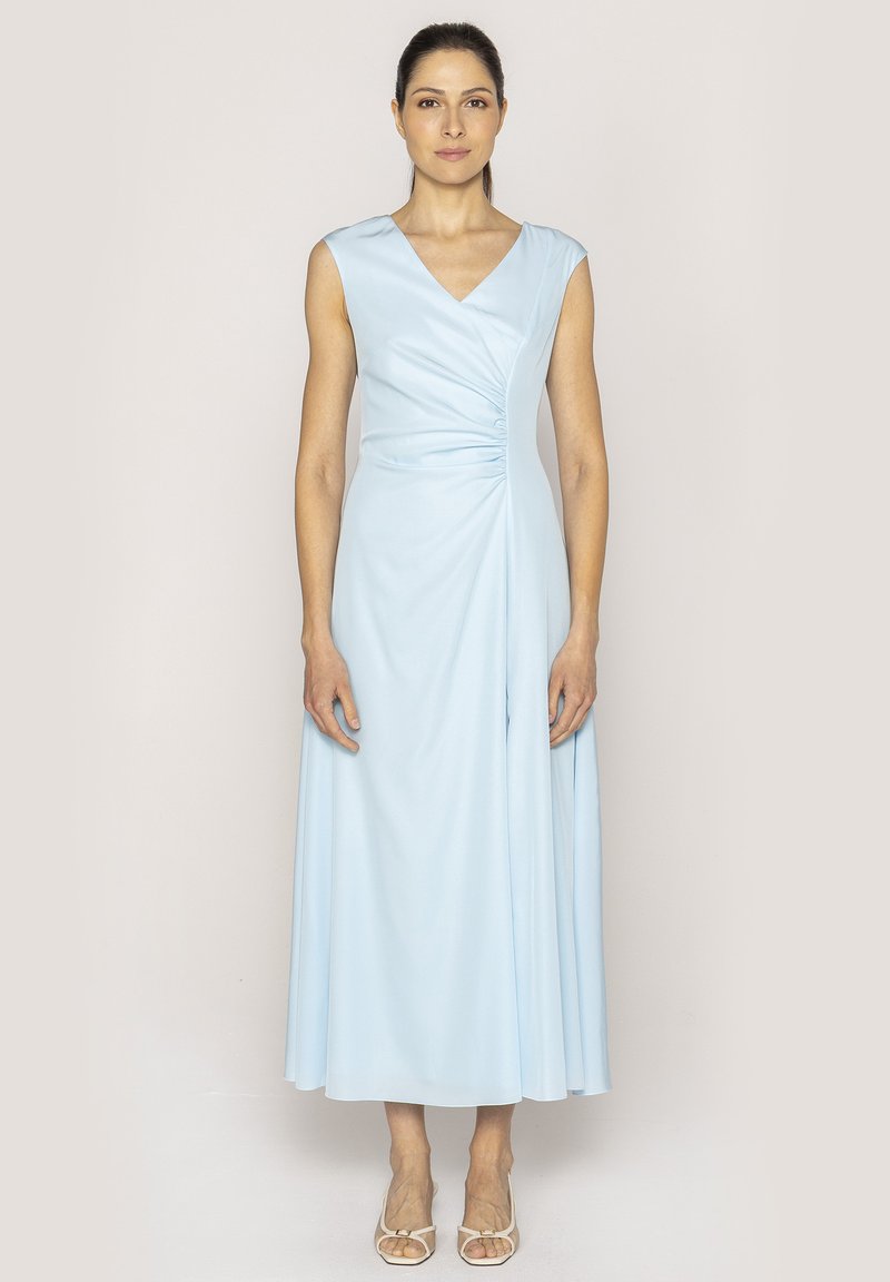 Jasnoniebieska sukienka bez rękawów z dekoltem w kształcie litery V, z drapowaną konstrukcją i marszczeniem w talii, sięgająca do długości midi. Pasujące sandały.
