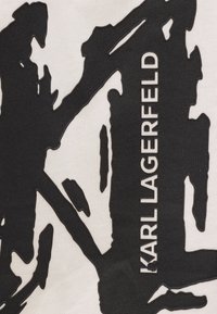 Juodai balta medvilnės medžiaga su ryškiu abstrakčiu raštu, apipjaustytais elementais ir tekstu "KARL LAGERFELD" didžiosiomis raidėmis.