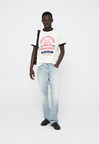 Λευκό μπλουζάκι με κόκκινο και μπλε γραφικό κείμενο, μαύρες λεπτομέρειες. Ανοιχτό μπλε τζιν, ελαφρώς φαρδύ στη βάση. Μαύρα παπούτσια. Απλός σχεδιασμός, casual στυλ.