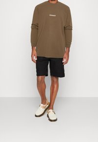 Ruskea pitkähihainen paita logolla, mustat shortsit ja beige sandaalit hihnoilla. Sileä pinta, rento tyyli ja väljä istuvuus.