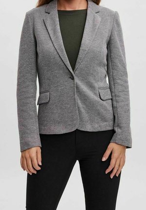 Femme portant un blazer gris ajusté sur un haut vert foncé et un pantalon noir, debout, les mains détendues le long du corps.