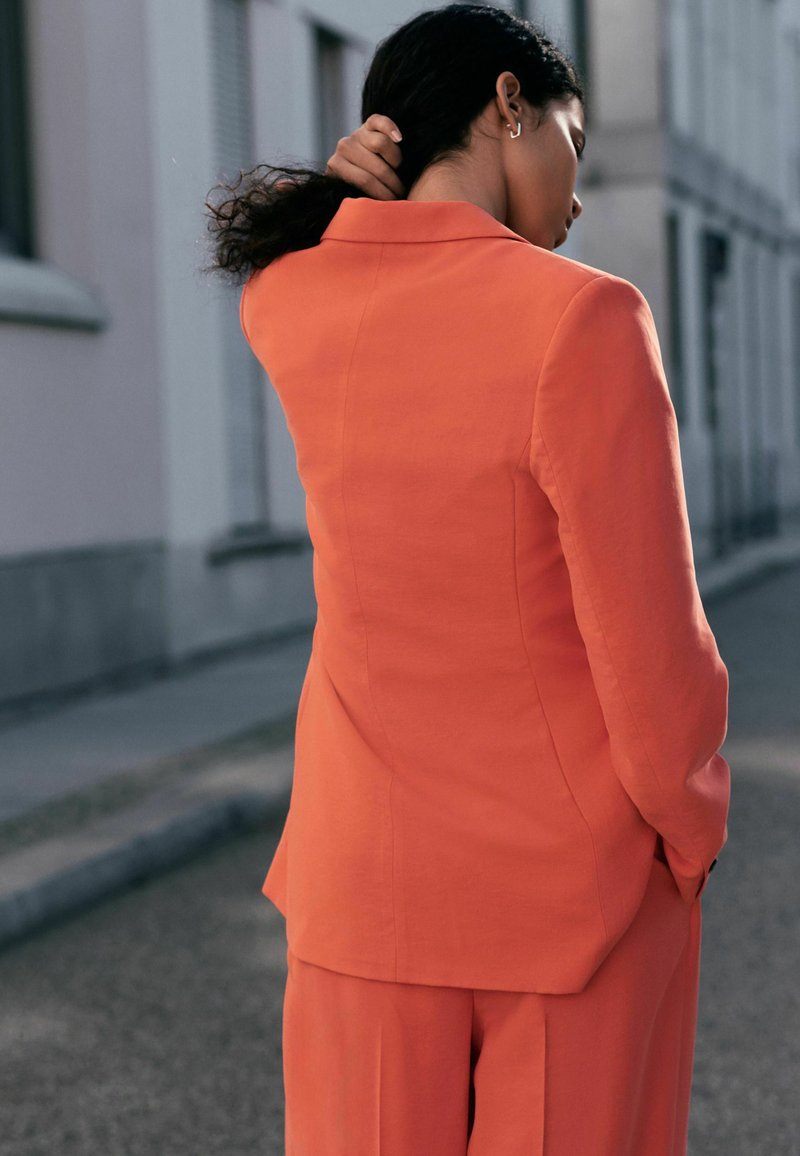 Donna con capelli ricci scuri che indossa un abito arancione, in piedi su una strada cittadina, si raccoglie i capelli in una coda bassa con una mano.