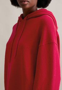 Felpa rossa con cappuccio caratterizzata da una texture morbida, vestibilità oversize e dettagli a coste sui polsini e sull'orlo. Cappuccio regolabile con coulisse.