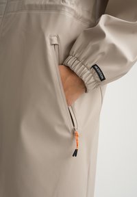 Béžový nepromokavý rukáv bundy s pružným lemem, viditelná ruka uvnitř boční kapsy na zip s oranžovým táhlem a černou štítkem „W.EDITION".