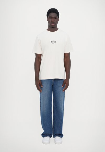 T-shirt blanche avec un logo noir, associée à un jean bleu ample et des baskets blanches. La tenue présente un design simple et décontracté.