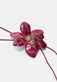 Ciondolo a forma di fiore viola con una finitura lucida, centro dorato intricatamente lavorato e un cordino rosso texturizzato per appenderlo.
