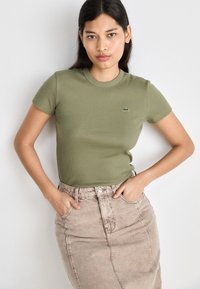 Olive green ribbed T-shirt with short sleeves, featuring a small embroidered logo. Paired with a light beige denim skirt.