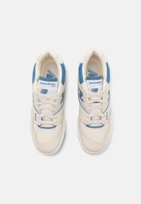 Béžové kožené tenisky s modrými akcenty, zaoblenou špičkou a plochými tkaničkami. Na jazyku a na boku se nachází logo "new balance" a číslo modelu.