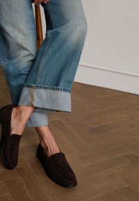 Brown suede loafers with stitching detail paired with light blue rolled hem jeans on a wooden floor. Simple and classic design.
