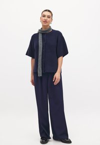 Marineblauwe gebreide cardigan met korte mouwen, ronde hals en knopen; gecombineerd met marineblauwe wijde broek en een grijze sjaal.