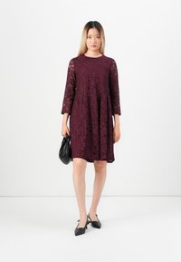 Femeie îmbrăcată într-o rochie dantelată bordeaux până la genunchi, cu mâneci lungi, ținând o geantă de mână neagră și purtând pantofi cu vârf ascuțit negri.