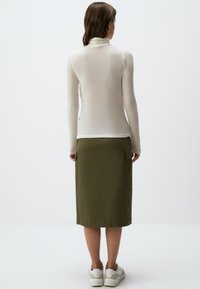 Maglione a collo alto color crema abbinato a una gonna verde oliva lunga fino al ginocchio. Dettagli notevoli includono una silhouette attillata e tasche laterali. Sneakers bianche.
