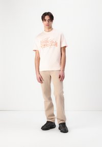 Camiseta de algodón rosa claro con un estampado gráfico en contraste, combinada con pantalones beige y zapatillas negras, exhibida sobre un fondo blanco.