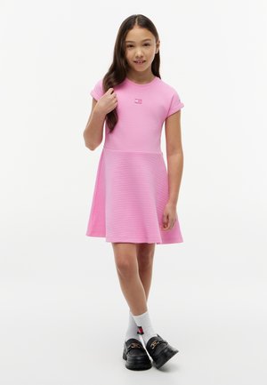 Mädchen mit langen dunklen Haaren, das ein rosa kurzärmeliges Kleid, weiße Socken und schwarze Loafers trägt, steht vor einem schlichten weißen Hintergrund.