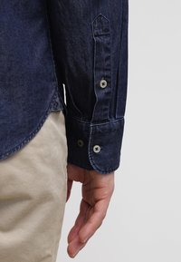 Gros plan sur la main d'une personne à côté d’un pantalon beige portant une manche de chemise en denim bleu foncé avec des poignets boutonnés.