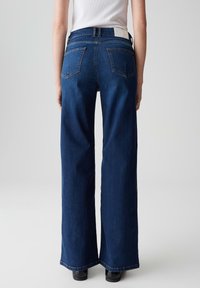 Persona che indossa jeans in denim a gamba larga di colore blu scuro e scarpe nere, vista di spalle su uno sfondo chiaro e semplice.