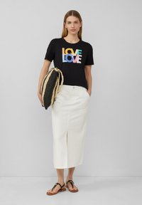 T-shirt noir à manches courtes avec un graphique coloré "LOVE", associé à une jupe midi blanche avec une fente frontale et des sandales brunes. Elle porte un sac rayé.