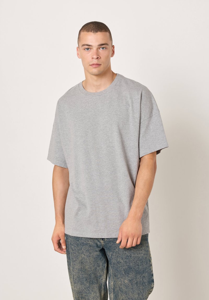 Šedé bavlněné tričko s kulatým výstřihem a krátkými rukávy, navržené pro volný střih. Nošeno s potrhanými modrými džínami.