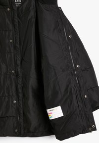 Schwarze gepolsterte Kinderjacke mit Reißverschluss und Druckknöpfen geöffnet, die das glatte Innenfutter und das Namensschild am inneren Saum zeigt.
