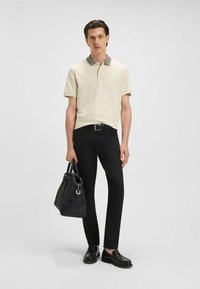 Beige polo shirt with a textured collar, paired with black trousers and slip-on black shoes. The model carries a black bag.