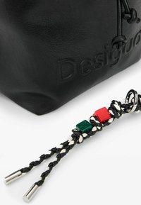 Black textured leather bag corner with embossed "Desigual" logo and attached black-and-white braided strap featuring red and green beads and metal rings.