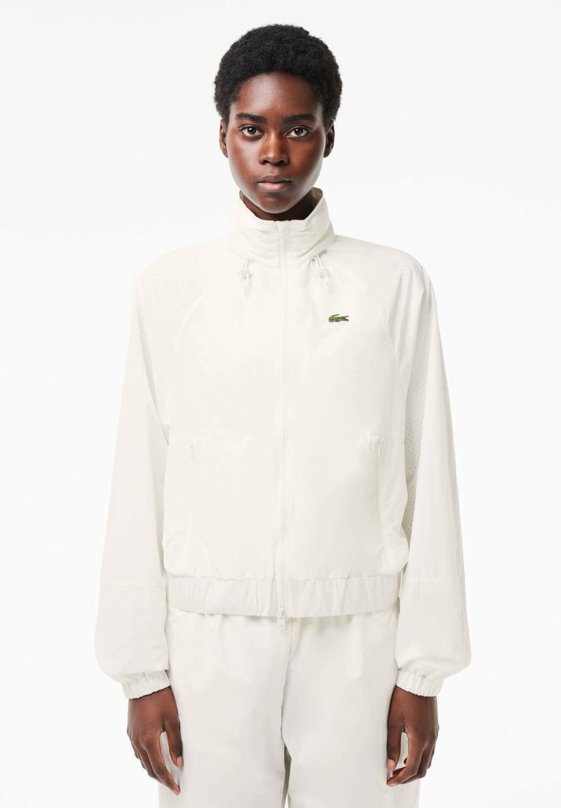 Osoba ubrana w białą kurtkę na zamek i dopasowane spodnie, stojąca na tle gładkiej białej ściany, zwrócona w stronę obiektywu.