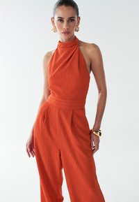 Oranžový overal na zavazování za krk s texturovanou látkou a plisovanými detaily. Má přiléhavý pas a široké nohavice. Zlaté doplňky.