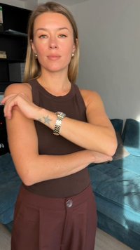 Brown sleeveless top and burgundy high-waisted trousers. Silver wristwatch with a patterned band. Background features a teal couch.