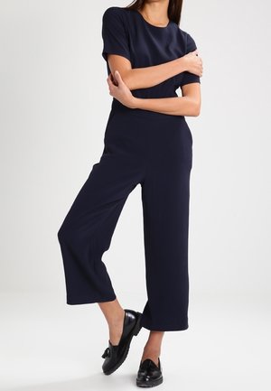 Marineblauwe jumpsuit met korte mouwen, wijde pijpen en een gladde textuur, gecombineerd met zwarte schoenen met een strikaccent.