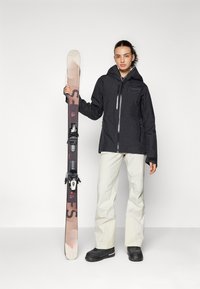 Schwarze wasserdichte Jacke mit Kapuze, cremefarbene Skihose und schwarze Skischuhe. Hält pinke und schwarze Skier mit Bindungen und Logo.