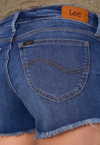 Shorts de mezclilla en azul oscuro. Presentan un dobladillo deshilachado, bolsillos traseros con costuras curvas y una etiqueta de cuero marrón en la cinturilla.