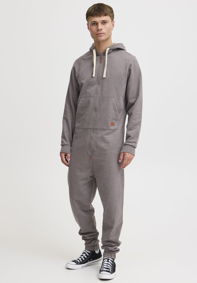 Gray zippered onesie with a hood, contrasting cream drawstrings, front pockets, and ribbed cuffs, paired with black sneakers.