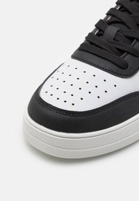 Schwarzer Ledersneaker mit weißer, perforierter Zehenkappe, strukturierten schwarzen Akzenten und weißer Gummisohle mit profiliertem Laufsohlenmuster.