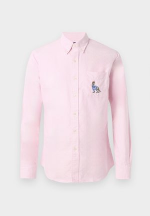 Camisa de manga larga de color rosa claro con cuello abotonado y diseño de perro bordado en el bolsillo izquierdo del pecho. Tejido de algodón, textura suave.