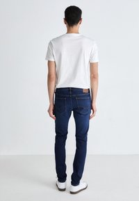 Jean en denim bleu foncé, coupe slim, avec des détails de couture subtils, associés à un T-shirt blanc simple et des baskets blanches.