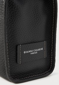 Melna ādas soma ar teksturētu apdari, kurai ir balta šuvumu un taisnstūra birka ar iespiestu uzrakstu "GIANNI CHIARINI FIRENZE."