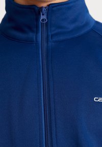 Suurennettu kuva tummansinisestä vetoketjulla varustetusta takin kauluksesta, jossa näkyy vetoketjun liukusoinnun osuus ja osittainen valkoinen logo oikealla puolella.