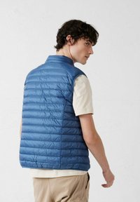 Chaleco acolchado azul con cuello alto y sin mangas; presenta costuras horizontales y un acabado de tela ligera y brillante.