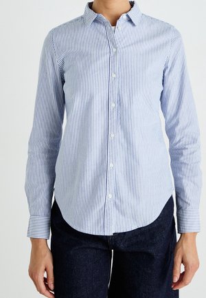 Person wearing a long-sleeve, light blue and white vertically striped button-up shirt and dark pants against a plain background.