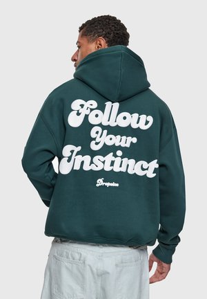 Mężczyzna w ciemnozielonej bluzie z kapturem na głowie, białych spodniach oraz wyraźnym białym napisie na plecach "Podążaj za swoim instynktem" i małym logo "Dropsize".