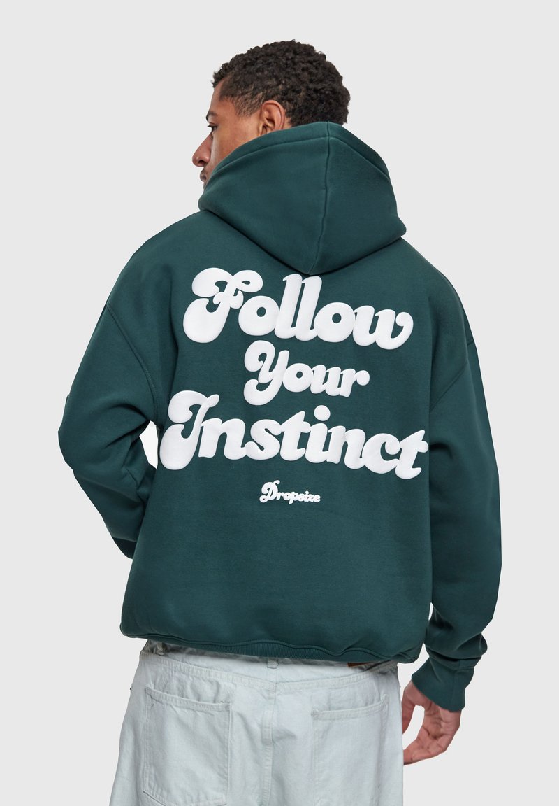 Homme portant un sweat à capuche vert foncé avec la capuche relevée, un pantalon blanc, et un texte blanc audacieux au dos indiquant "Suis ton instinct" avec un petit logo "Dropsize".