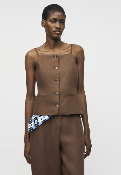 Γυναίκα που φορά καφέ αμάνικο τοπ με κουμπιά και ταιριαστό παντελόνι, με μπλε και λευκό υφασμάτινο μοτίβο που φαίνεται, όρθια μπροστά σε λευκό φόντο.