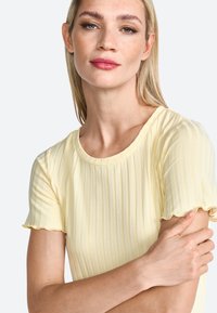 Top a coste giallo chiaro con scollatura rotonda e maniche corte a volant. La texture morbida e il design aderente esaltano il suo aspetto elegante.